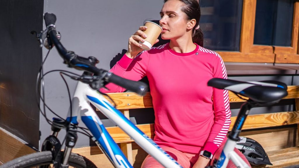 Woman in pink sweatshirt resting with a coffee by bicycle outdoors.